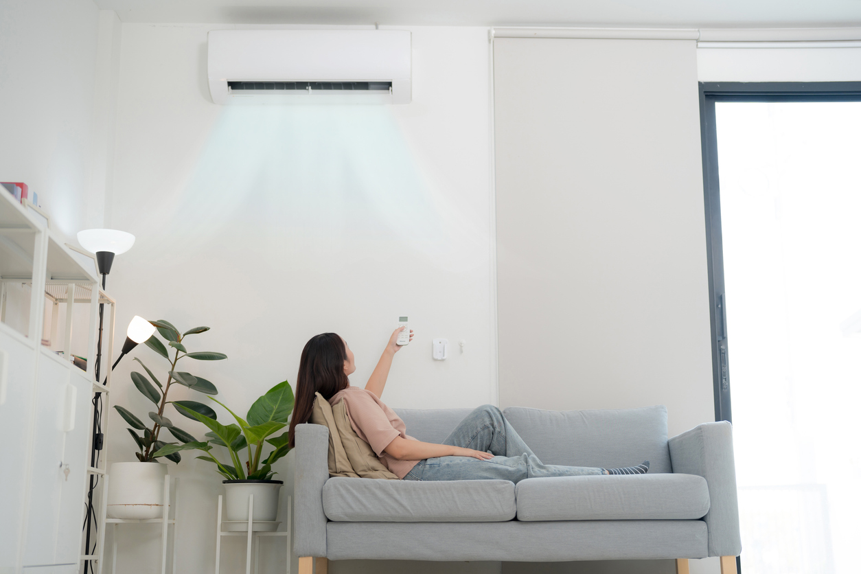 Woman relaxing on a sofa using a remote to operate an air conditioner at home, representing concerns about strange or bad AC smells.